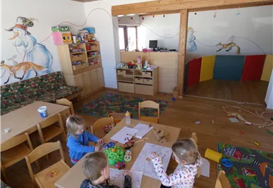 A colorful playroom for children with tables, chairs, and a variety of craft materials. Bright drawings adorn the walls, along with a play area featuring colorful partitions.