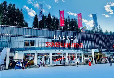 A modern ski shop with the sign "HANSIS SKIVERLEIH" in the midst of a snowy environment. In the background, trees and a blue sky can be seen.