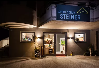 Ein sportlicher Schuhladen mit dem Namen „Sport Schuh Steiner“ ist bei Nacht beleuchtet. Der Eingangsbereich ist einladend, mit dekorativen Elementen an der Fassade.
