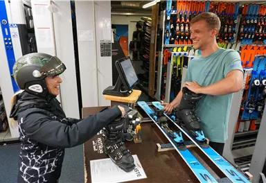 A female skier receives her equipment in a sports store. The staff member kindly helps her with putting on her ski boots.