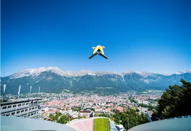Ein Skispringer springt über eine malerische Stadt und die umliegenden Berge. Der Himmel ist klar und blau.
