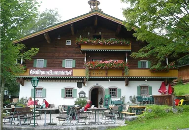 A traditional wooden house with blooming balconies and an inviting terrace. Guests sit outside and enjoy the peaceful surroundings.