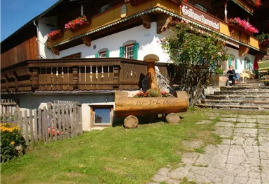 A traditional Alpine house with balconies and flower boxes. In the foreground, there is a wooden bench and the path leads to the stairs.