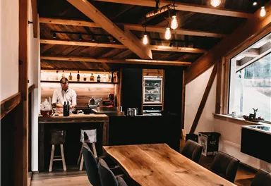 Ein gemütlicher Raum mit einer großen Holztisch und gepolsterten Stühlen. Im Hintergrund ist eine offene Küche mit warmen Holzdetails und sanfter Beleuchtung zu sehen.