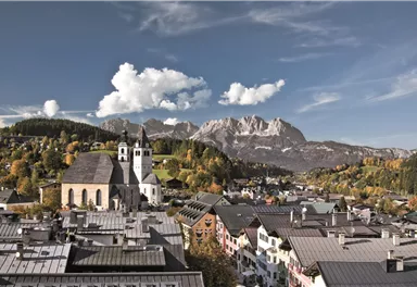 Eine malerische Stadt mit einem historischen Kirchturm und Bergen im Hintergrund. Die Landschaft ist von bunten Bäumen umgeben und der Himmel ist teilweise bewölkt.