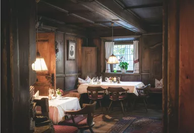 Ein gemütlicher Speisesaal mit hölzernen Wänden und warmem Licht. Tische sind eingedeckt und ein Fenster mit Blick auf Pflanzen sorgt für eine einladende Atmosphäre.