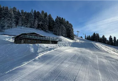 A winter landscape with a house on a snowy slope. In the background, trees and a ski lift are visible.