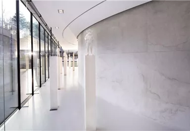 Ein moderner Innenraum mit großen Fenstern und einer hellen, minimalistischen Gestaltung. Auf weißen Sockeln stehen Skulpturen, die den Raum ausfüllen.