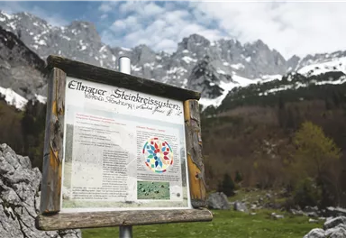 An informational sign about the Ellmauer Stone Circle systems in a mountainous landscape. Snow-capped peaks and forested slopes are visible in the background.