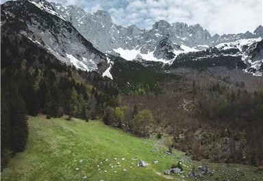 An impressive mountain landscape with snow-capped peaks and a green meadow. The area is surrounded by forests and has a cloudy sky.