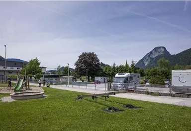 Ein ruhiger Campingplatz mit Wohnmobilen und einer Spielecke. Im Hintergrund sind grüne Wiesen und Berge zu sehen.