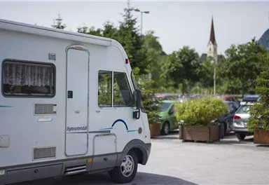 Ein Wohnmobil steht auf einem Parkplatz, umgeben von Autos und Bäumen. Im Hintergrund ist ein Kirchturm zu sehen.
