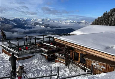 A mountain cabin surrounded by snow with a view of the mountains. The atmosphere is clear and sunny.