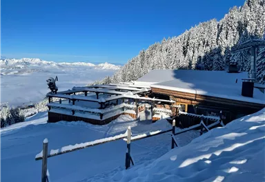 A snowy mountain house surrounded by snow-covered trees. The sky is clear and blue, with a view of the mountains in the distance.