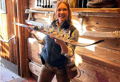 A cheerful woman in traditional Bavarian clothing is holding a ski boot with glasses in her hand. In the background, there are wine bottles and a rustic wooden paneling visible.