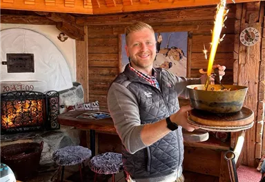 A cheerful waiter carries a dish with sparkling sparks in a paneled wooden restaurant. In the background, a cozy atmosphere is visible with a fireplace and wooden chair.