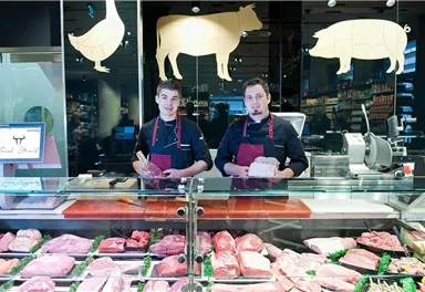 Two butchers stand behind a counter with fresh meat. The display shows a variety of meat types in a modern butcher shop.