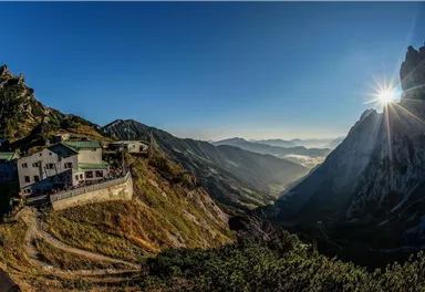 An impressive mountain landscape with an inn on a terrace. The sun rises on the horizon and illuminates the valley.