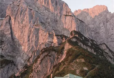 An impressive mountain landscape with steep cliffs and a cabin building in the foreground. The cliffs are illuminated in soft evening light tones.