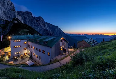 A picturesque mountain hotel at dusk, surrounded by majestic mountains. The windows emit warm light, creating an inviting atmosphere.