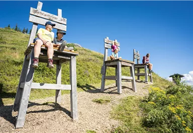 Große Holzstühle auf einem hügeligen Gelände. Menschen sitzen darauf und genießen die Aussicht bei sonnigem Wetter.