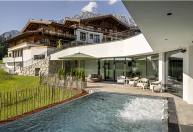 Ein modernes Haus mit großen Fenstern und einem angrenzenden Pool. Im Hintergrund sind grüne Wiesen und Berge zu sehen.