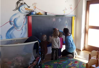 A bright classroom with a blackboard where one person and two children are drawing. A cheerful snowman is painted on the wall.