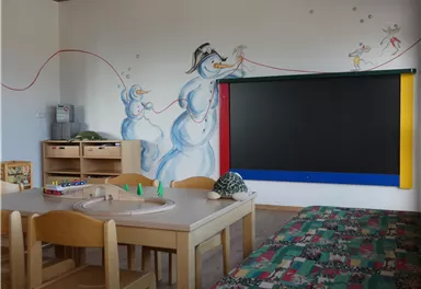 A cheerful room with a table and chairs, ideal for children. Funny snowmen are painted on the wall, and there is a large black board.