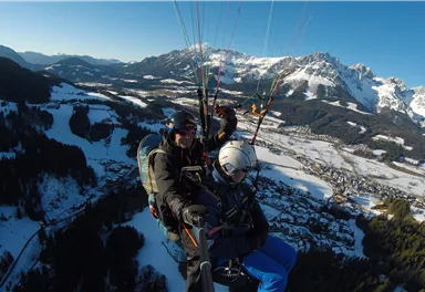 Ein Tandemflug mit Paragliding über schneebedeckte Berge. Im Hintergrund sind malerische Täler und eine klare, blaue Himmel zu sehen.