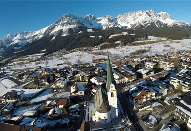 Eine malerische Berglandschaft mit schneebedeckten Gipfeln und einem charmanten Dorf im Vordergrund. Die Kirche mit dem hohen Turm ist ein zentraler Blickfang.