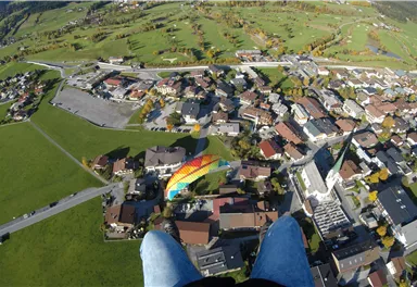 Eine Luftaufnahme einer Stadt mit grünen Wiesen und Wohnhäusern. Im Hintergrund sind Berge und ein bunter Gleitschirm zu sehen.