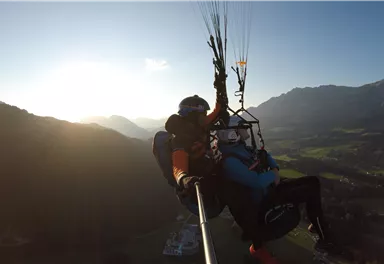 Ein Mensch schwebt mit einem Gleitschirm über einer malerischen Landschaft. Die Sonne scheint und die Berge sind im Hintergrund sichtbar.