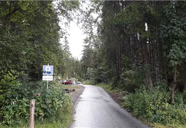 Ein schmaler Weg führt durch einen Wald mit hohen Bäumen. Links ist ein Schild sichtbar, und im Hintergrund parken einige Fahrzeuge.