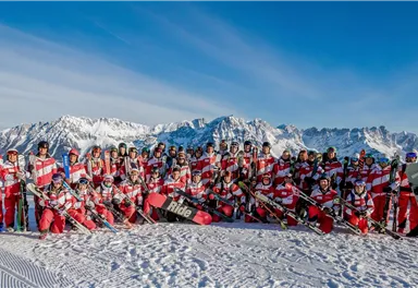A large group of skiers in red and white suits stands on a snow-covered mountain. In the background, impressive mountains and a blue sky can be seen.