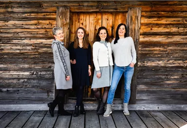 Four women are happily standing in front of a rustic wooden wall. They are wearing various stylish outfits and have different hairstyles.