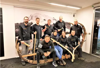 Eine Gruppe von neun Personen posiert fröhlich in einem Raum. Sie stehen um einen traditionellen Schlitten aus Holz und tragen schwarze Jacken.