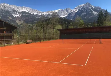 Ein Tennisspielplatz mit rotem Belag, umgeben von majestätischen Bergen und einer klaren blauen Himmel. Die Natur fügt eine malerische Kulisse hinzu.