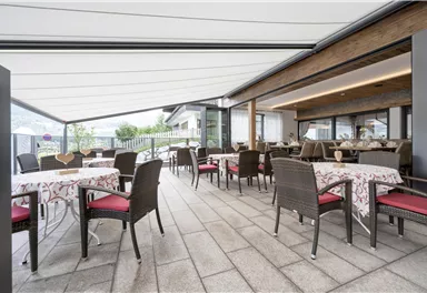 Ein modernes Restaurant mit stilvollen Tischen und Stühlen auf einer überdachten Terrasse. Die Umgebung ist hell und einladend, ideal für Mahlzeiten im Freien.
