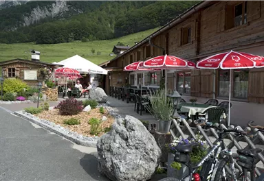 Ein gemütliches Restaurant mit Holzverkleidung, Terrassenstühlen und roten Sonnenschirmen. Im Hintergrund sind grüne Hügel und Bäume zu sehen.