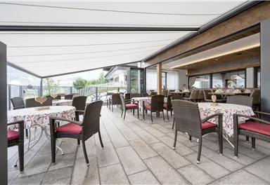 Ein modernes Restaurant mit stilvollen Tischen und Stühlen auf einer überdachten Terrasse. Die Umgebung ist hell und einladend, ideal für Mahlzeiten im Freien.