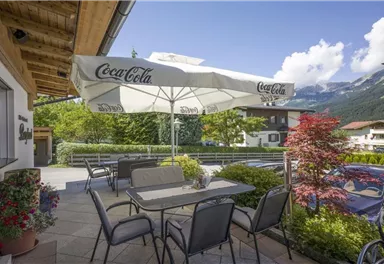 A cozy terrace with a large sunshade and seating. In the background, green trees and mountains can be seen.