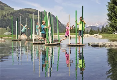 Kinder spielen auf schwimmenden Plattformen in einem klaren See. Im Hintergrund sind grüne Wiesen und Berge zu sehen.