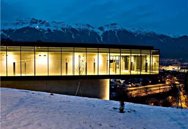 Ein modernes Gebäude mit großen Glasfenstern steht auf einem Hügel im Schnee. Im Hintergrund sind schneebedeckte Berge und ein Nachtblick auf die Stadt zu sehen.