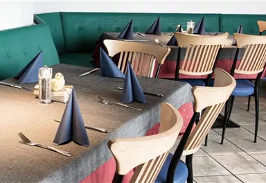 Ein gemütliches Restaurant mit Tischen, die mit grauen Tischtüchern und blauen Servietten dekoriert sind. Die Sitzgelegenheiten umfassen Holzstühle und eine gepolsterte Bank.