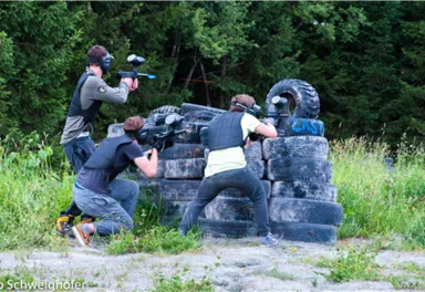 Three people are playing in an outdoor area with paintball markers. They are taking cover behind a pile of tires and are surrounded by trees.