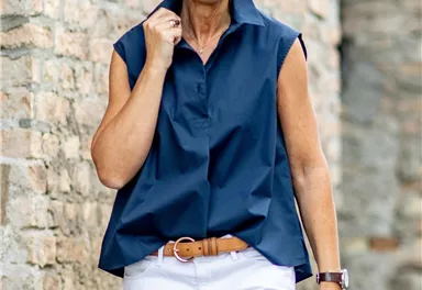 A smiling woman is wearing an elegant, sleeveless, blue shirt and white pants. She is standing in front of an old stone wall.