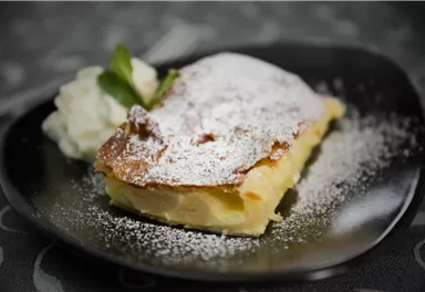 A piece of apple strudel on a black plate, sprinkled with powdered sugar. Next to it, a serving of cream and a mint leaf.