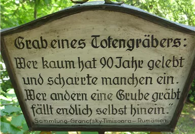 Ein Schriftzug auf einer Holztafel im Freien. Der Text thematisiert das Leben und den Tod mit einem nachdenklichen Spruch.