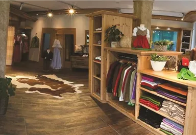 Ein gemütliches Geschäft mit traditioneller Kleidung und Holzregalen. Im Hintergrund sieht man eine Treppe und ein einladendes Ambiente.