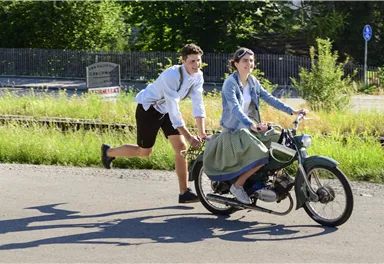 A boy is pushing a girl who is sitting on a motorcycle while they ride on a street. It is a sunny day with lots of green in the background.